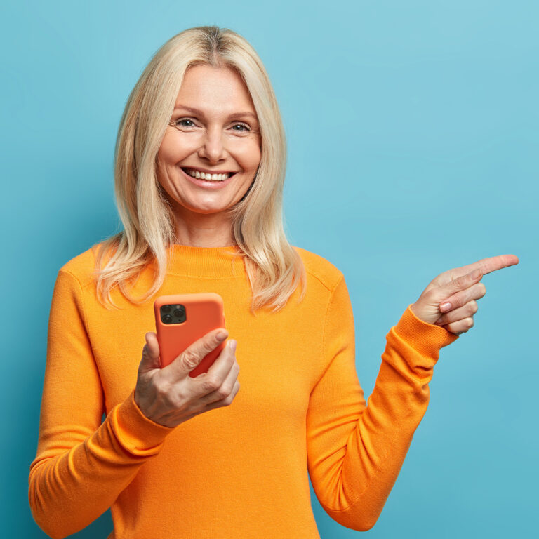 Positive blonde woman has happy smile holds mobile phone points away on copy space uses modern technologies being always in touch isolated on blue background. Middle aged lady with cellular.
